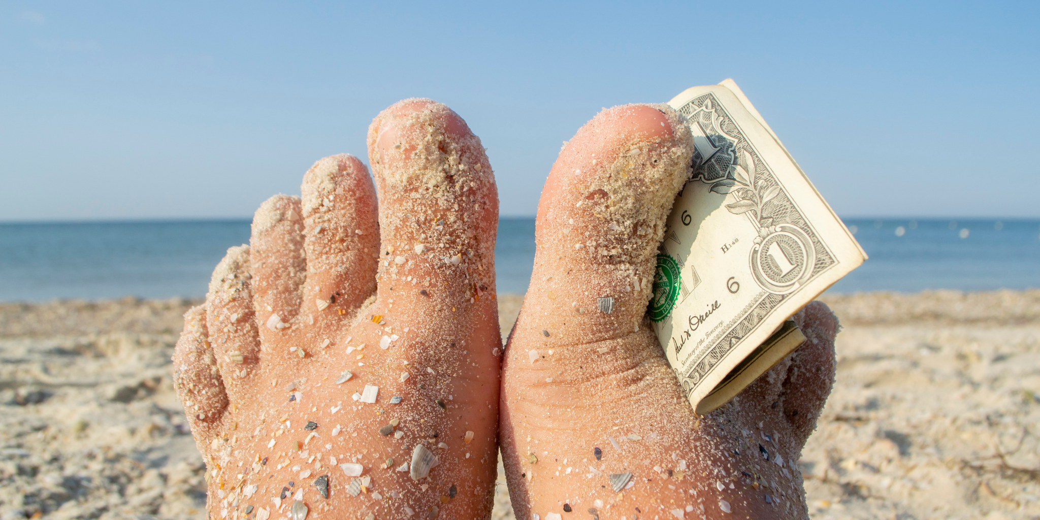 Feet in beach sand with a dollar bill tucked between the toes
